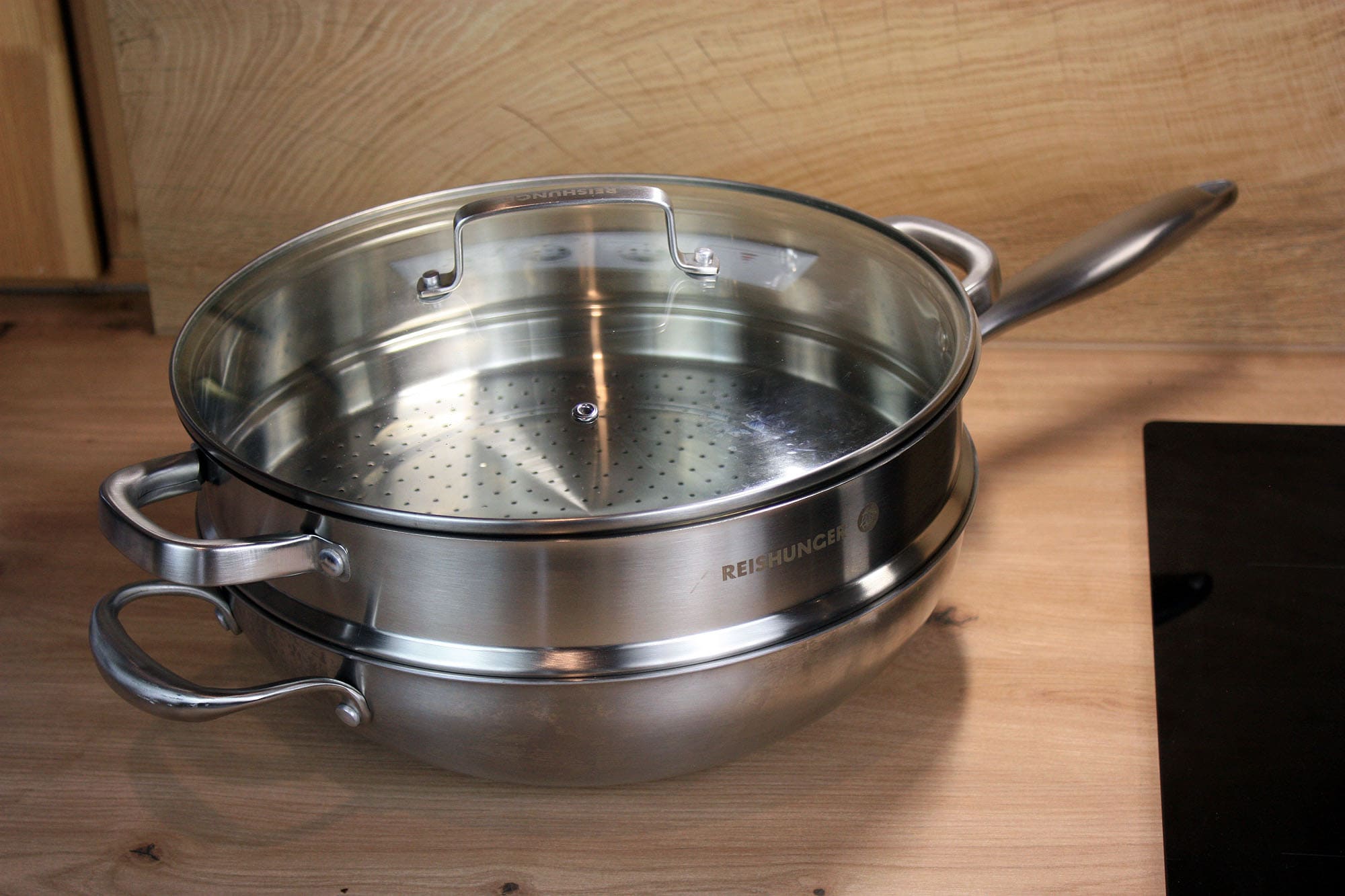 Die beste Wokpfanne 170 Edelstahl-Wok mit eingesetztem Dämpfeinsatz und aufgelegtem Glasdeckel auf einer Arbeitsfläche.