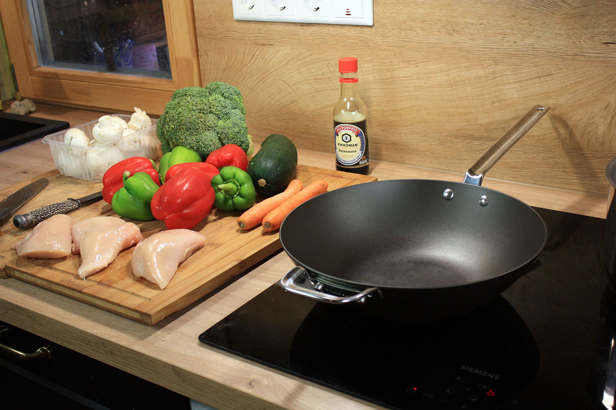 Die beste Wokpfanne 183 SCANPAN Black Iron 30 cm auf einem Induktionskochfeld neben Gemüse, Möhren, Brokkoli und rohem Hähnchenfleisch.